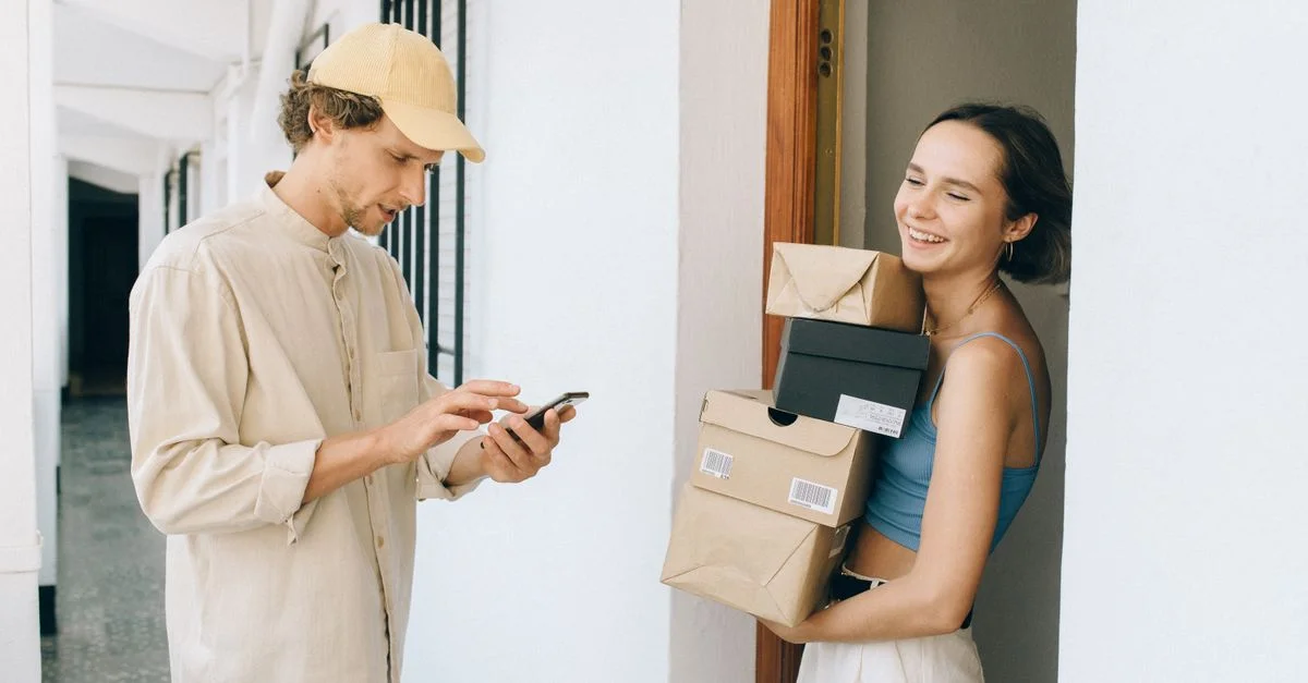 Une femme joyeuse reçoit une livraison d'une personne dans un couloir, utilisant un smartphone pour un paiement en ligne.